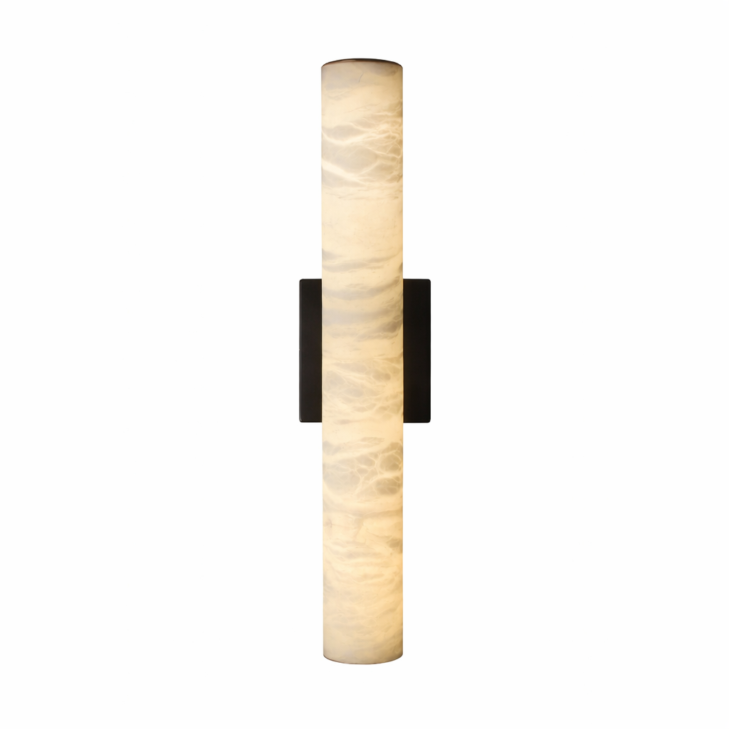 The slender cylindrical silhouette of the Velato marble wall light casting a soft, elegant glow against a contemporary textured backdrop.