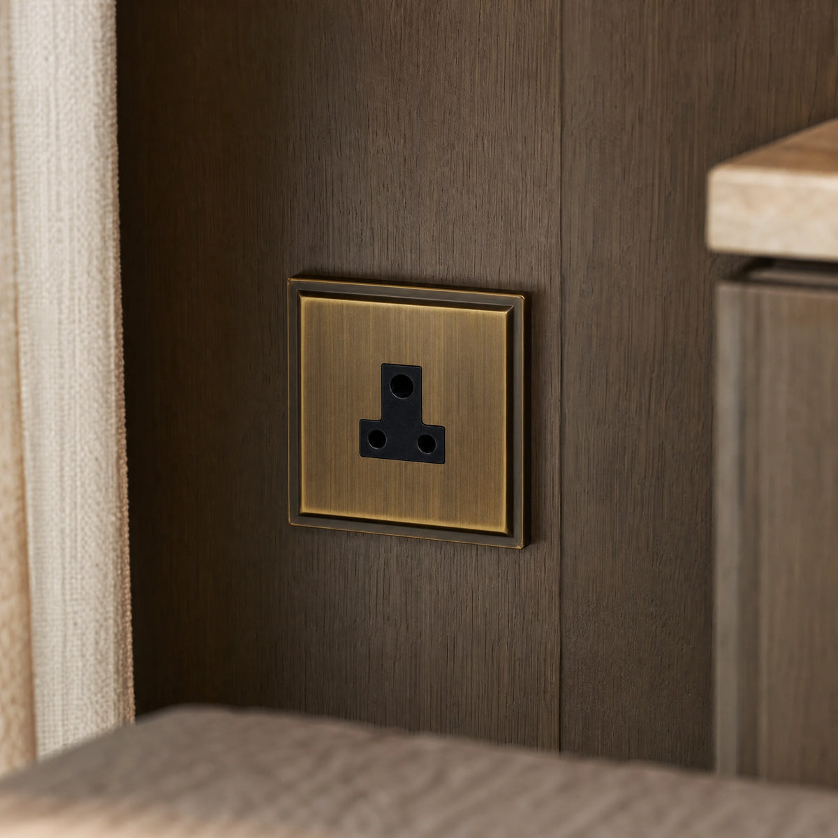Close-up of a luxury antique brass 5a socket installed within bespoke bedside joinery or low-level wall beside a luxury seating area by VORELLI®.