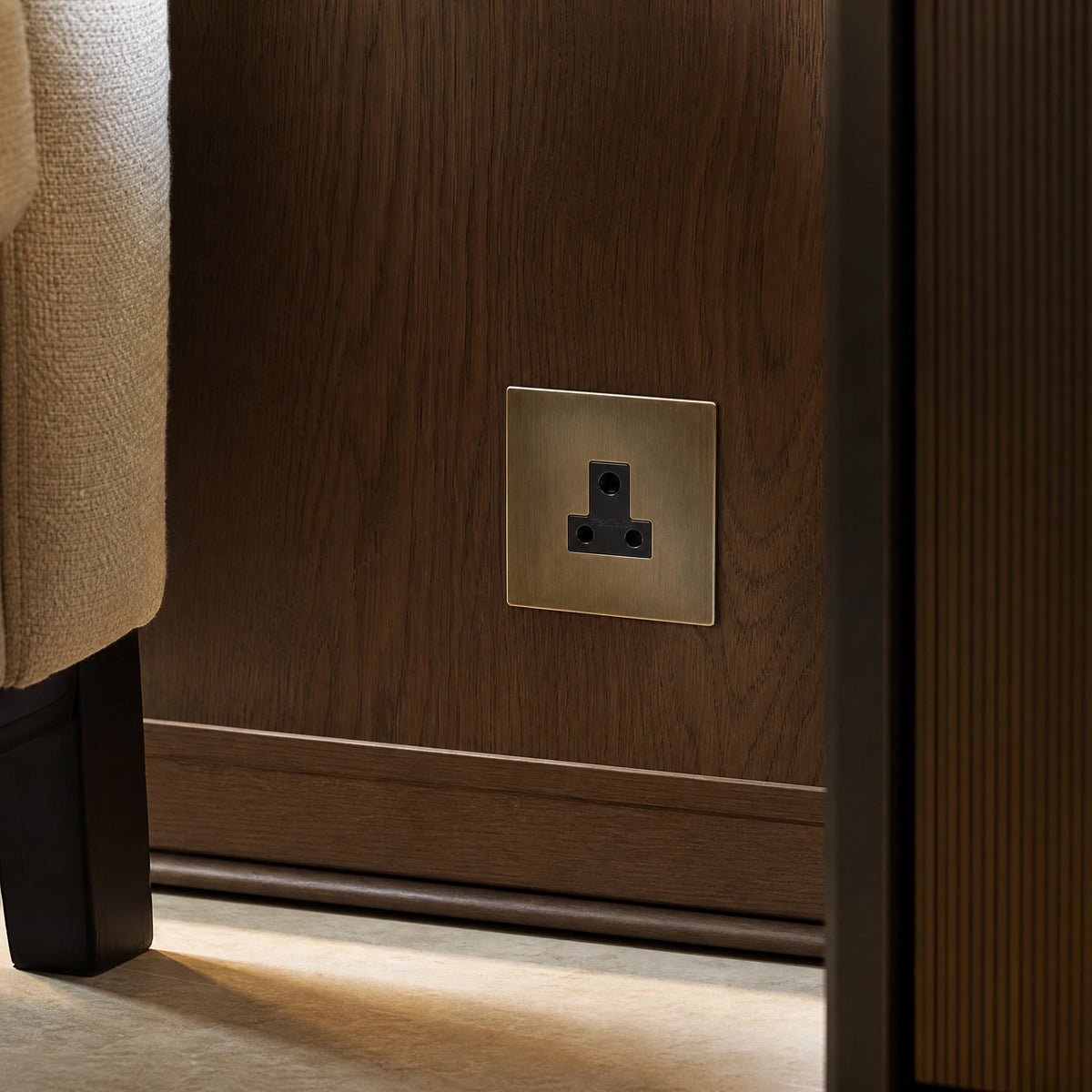 Close-up of a luxury antique brass 5a socket installed within bespoke bedside joinery or low-level wall beside a luxury seating area by VORELLI®.