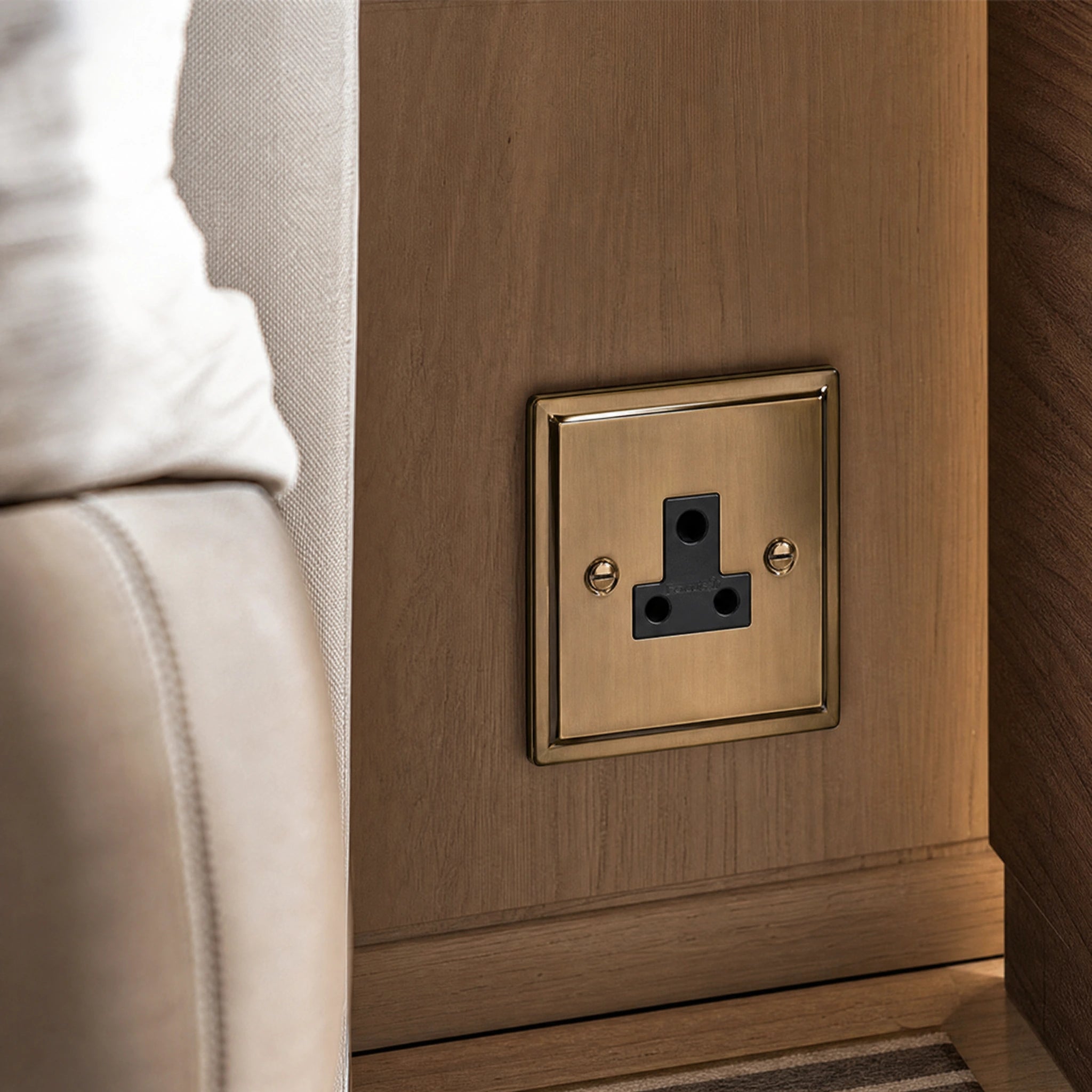 Close-up of a luxury antique brass 5a socket installed within bespoke bedside joinery or low-level wall beside a luxury seating area by VORELLI®.