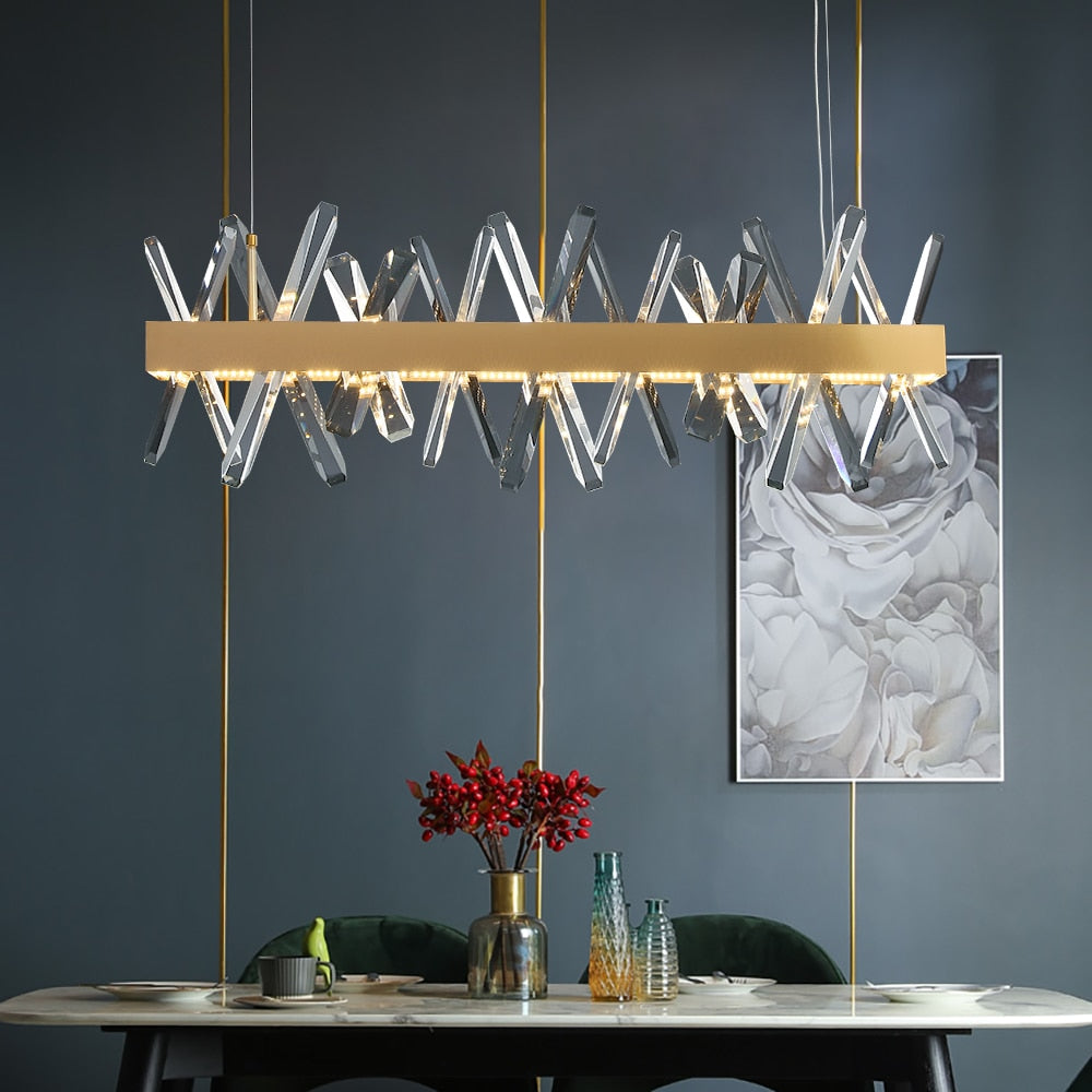 A contemporary rectangular chandelier with angled diamond cut crystals and a sleek horizontal stainless steel frame, hanging over a modern dining table with decorative flowers and glasses.